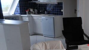 Eine kleine Küche mit weißen Schränken, einer Mikrowelle und einem Spülbecken. Ein schwarzer Stuhl und ein kleiner Tisch mit einem roten Gegenstand darauf.