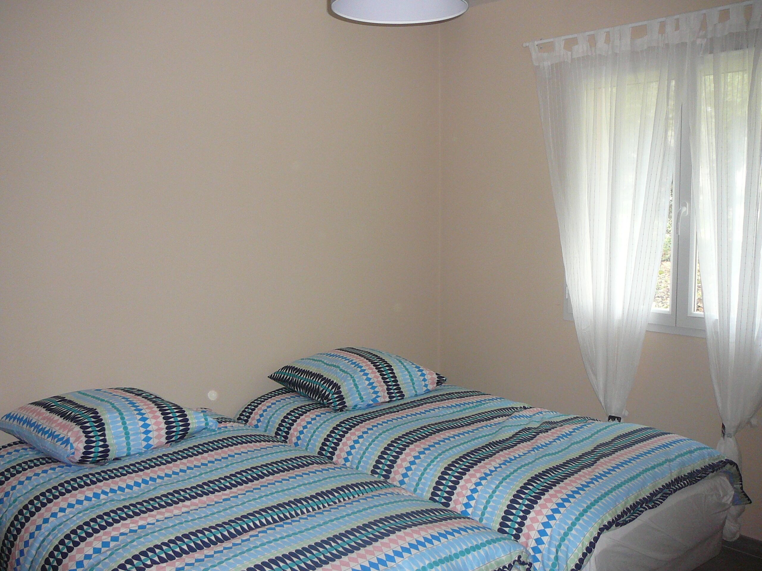 A bedroom with a bed covered in a patterned bedspread, a window with sheer curtains, and a white ceiling light.