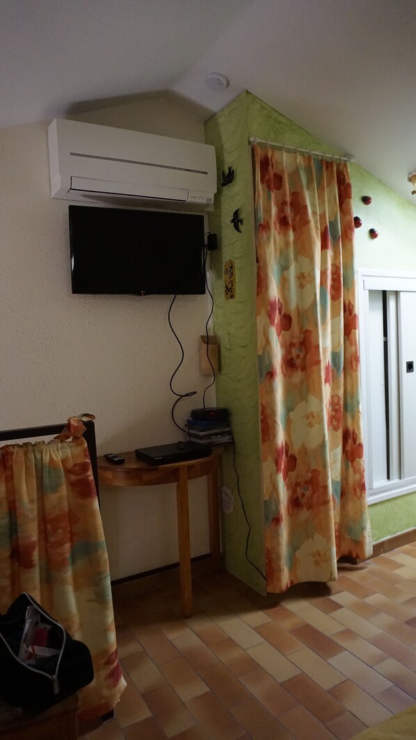 Una habitación con televisión de pantalla plana montada en la pared, una mesa de madera con algunos objetos, una pared verde con detalles decorativos y una cortina estampada.