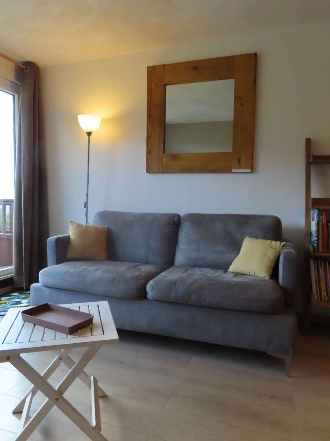 Un salon avec un canapé gris, deux coussins, un miroir à cadre en bois, une lampe sur pied et une petite table avec un plateau.