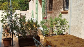 Eine Terrasse mit Tisch und Stühlen, umgeben von Blumentöpfen und einem Gebäude mit grünen Fensterläden.