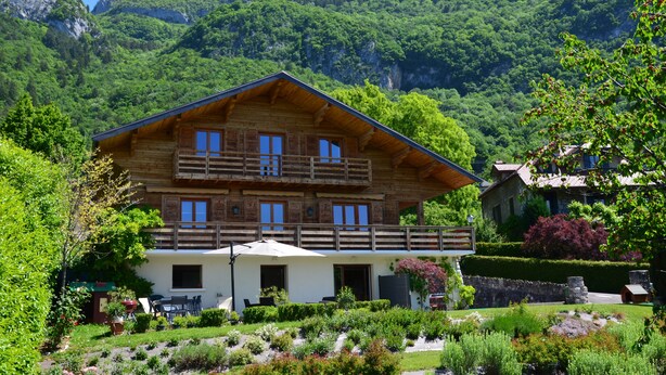 Eine Holzchalet mit einem Balkon, umgeben von üppiger Vegetation und Bergen im Hintergrund.