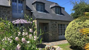 A stone house with a sloped roof, large windows, and a well-maintained garden with flowering plants and a neatly trimmed bush.