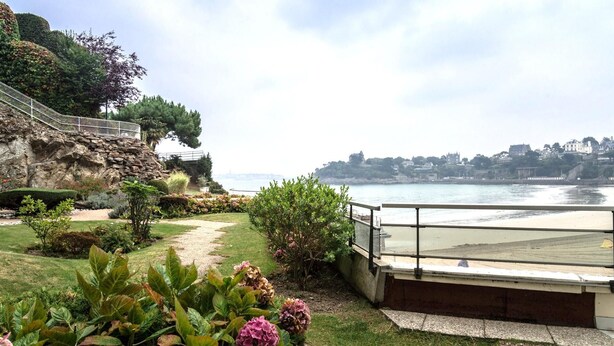 A coastal garden with a stone wall, a paved path, and a view of the sea and distant buildings.