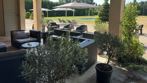 Um pátio externo com móveis de vime, guarda-sóis e vista para uma piscina e árvores.