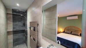 Casa de banho com chuveiro, sanita e lavatório. Espelho reflete um quarto com cama e mesa de cabeceira.