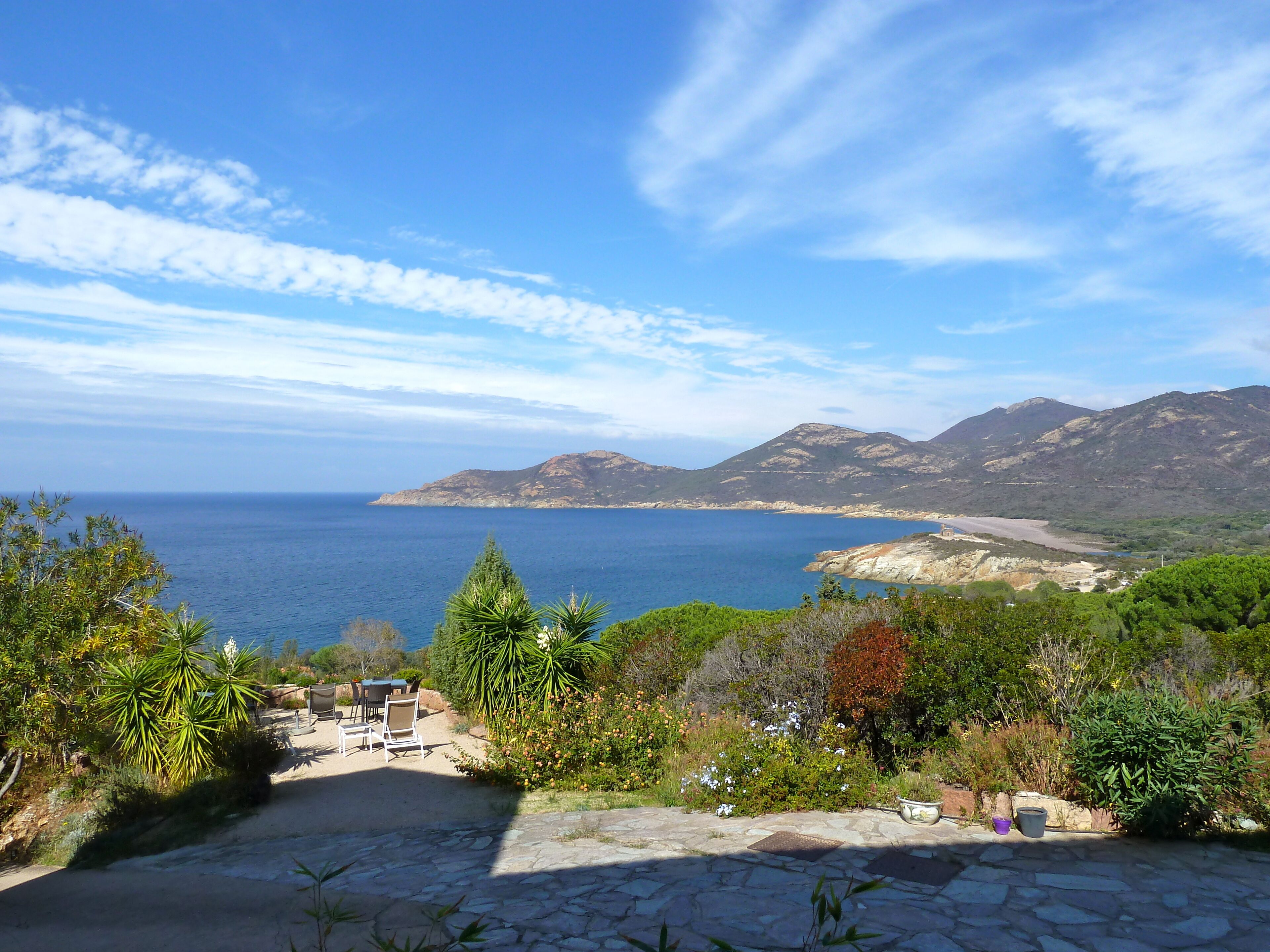 A stone patio with outdoor furniture, overlooking a coastal landscape with mountains and the sea.