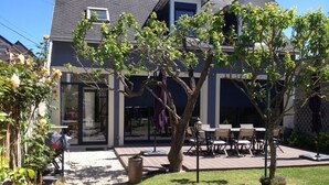 Ein zweigeschossiges Haus mit einer Terrasse, Sitzgelegenheiten im Freien und einem Garten mit Blumen.