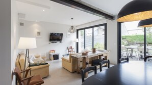 Ein modernes Wohnzimmer mit einer Couch, einem Esstisch und Stühlen. Ein Fernseher ist an der Wand befestigt und ein großes Fenster sorgt für natürliches Licht.