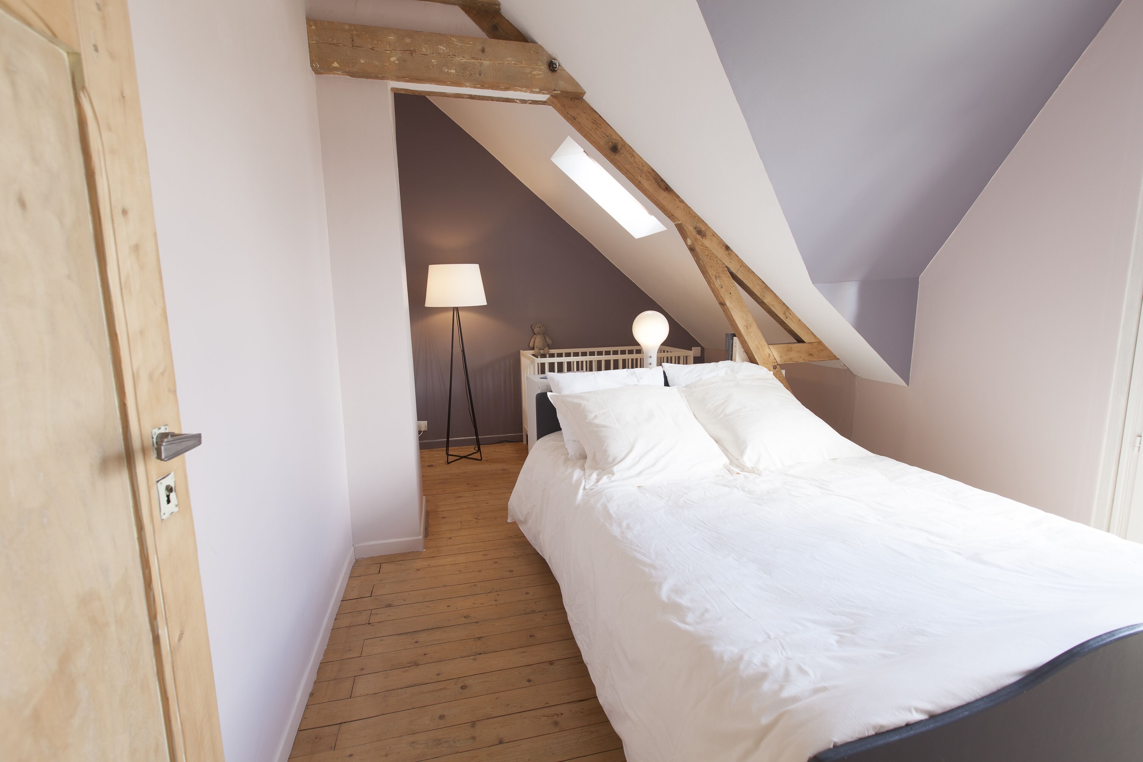 Une chambre avec un plafond en pente, un plancher en bois, un lit avec une literie blanche, une lampe de chevet et une petite table avec une lampe.