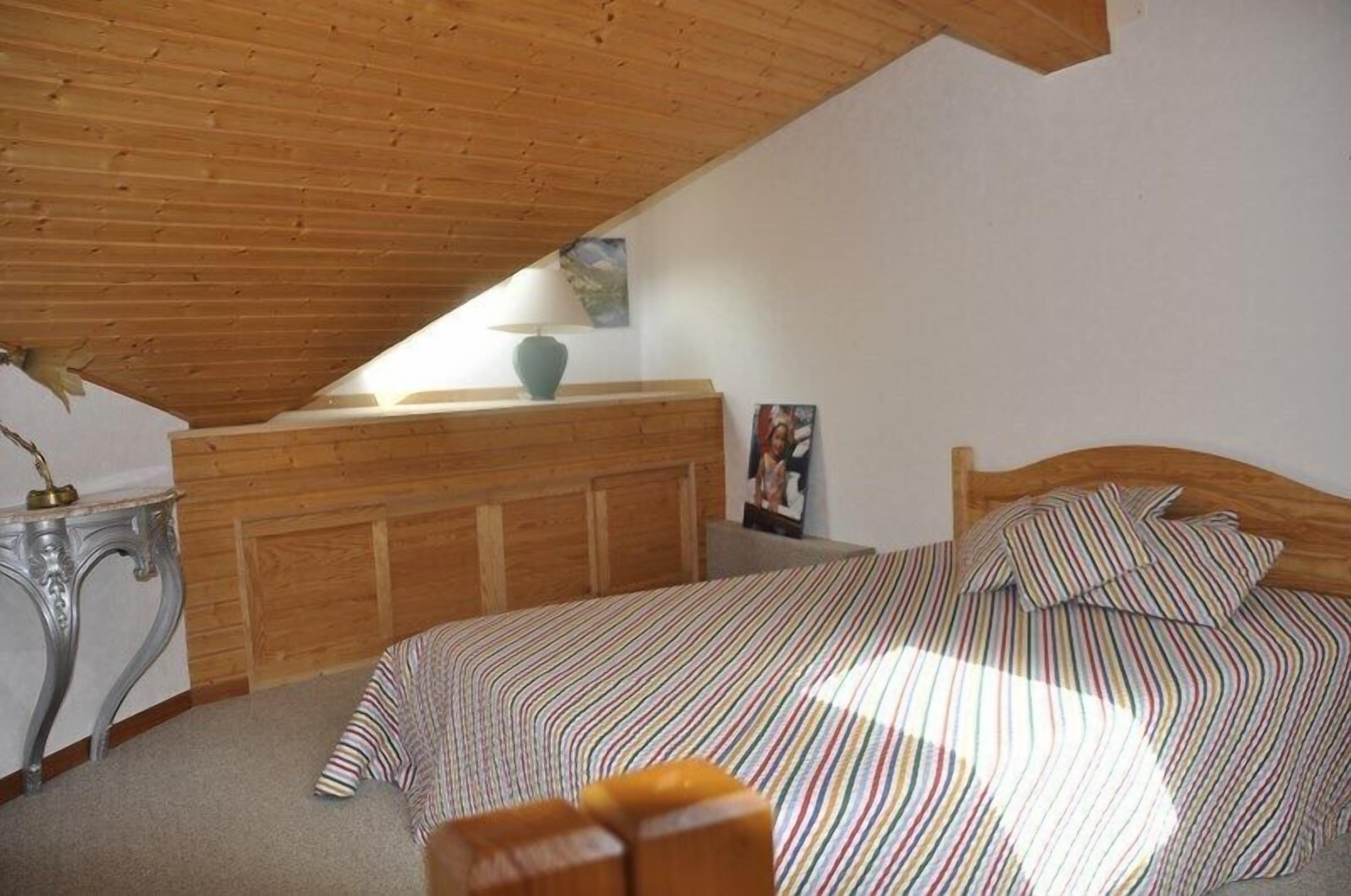Quarto com teto inclinado, cabeceira de madeira, colcha listrada, mesa de cabeceira com abajur e quadro na parede.