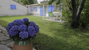 En trädgård med en krukväxt med lila blommor, en stenlagd gång, en vit byggnad med blåa fönsterluckor och ett bord med stolar.