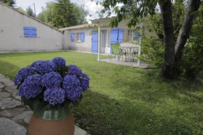 Un jardín con una planta en maceta de flores moradas, un sendero de piedra, un edificio blanco con contraventanas azules y una mesa con sillas.