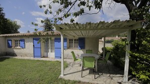 En täckt uteplats med ett bord och stolar, en pergola och ett hus med blåa fönsterluckor.
