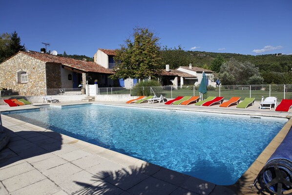 Una piscina con sillones de descanso, un edificio de piedra y un área cercada con árboles al fondo.