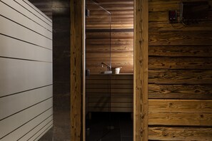 Interieur van een houten blokhut met een houten wand, een metalen plank met een witte container, en een klein elektronisch apparaat aan de wand.