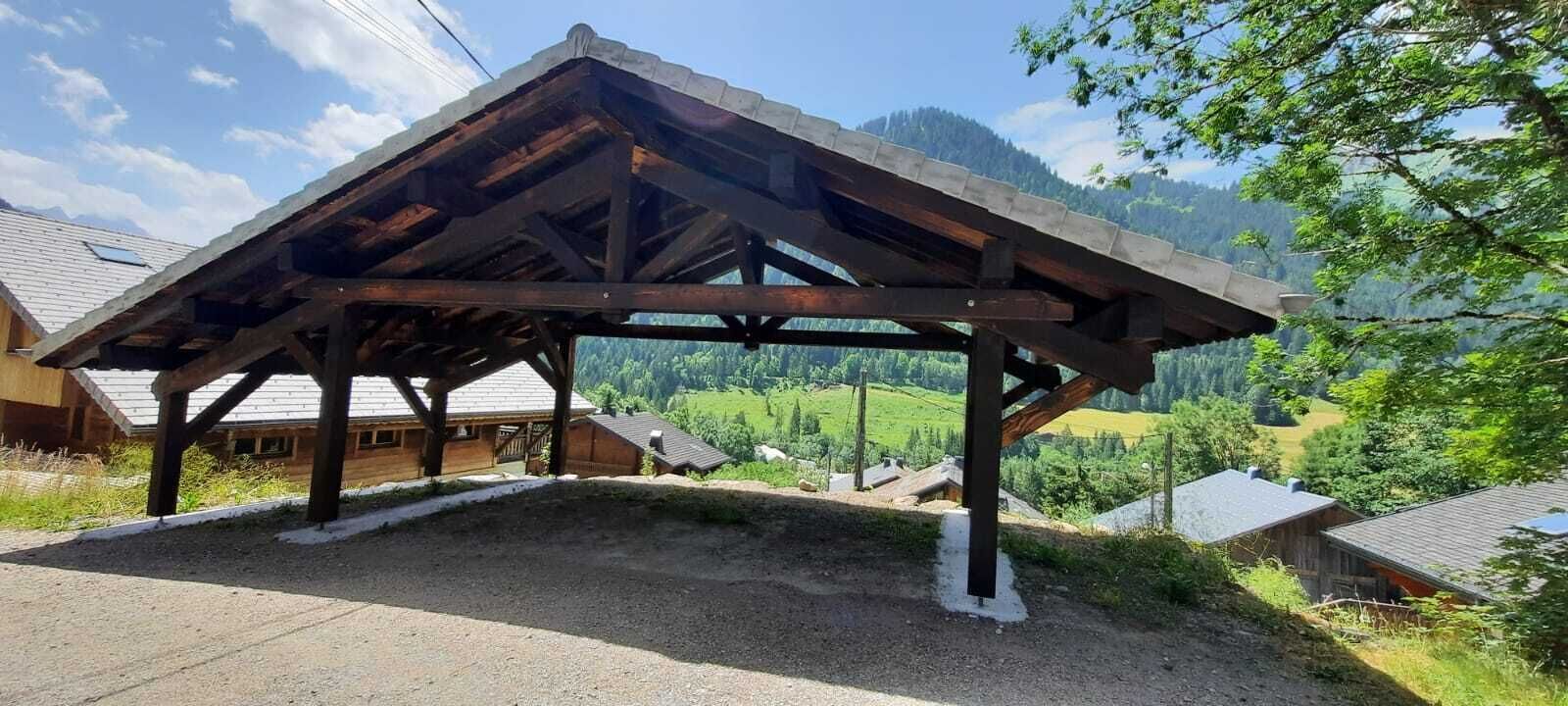 A wooden pavilion with a sloped roof, surrounded by a rural landscape with houses and mountains in the background.
