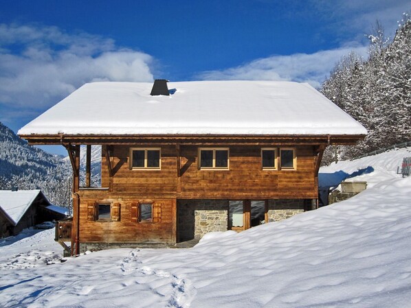 Un chalet en bois, dont le toit est recouvert de neige, situé sur une colline enneigée avec des montagnes en arrière-plan.