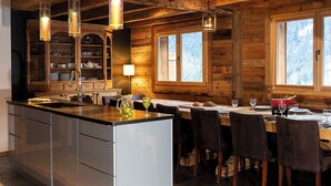 Interieur van een houten chalet met een keuken en eetgedeelte, met een groot eiland, houten kastjes en een raam met uitzicht op besneeuwde bomen.
