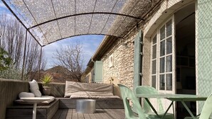 An outdoor patio with a wooden deck, a covered seating area with cushions, a metal table, and plastic chairs. - Stone farmhouse with swimming pool between Avignon and Orange, in Roquemaure (Roquemaure)