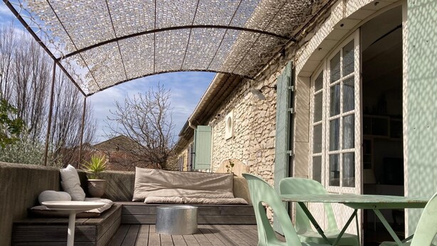 Una terrazza esterna con un pavimento in legno, una zona salotto coperta con cuscini, un tavolo in metallo e sedie di plastica.