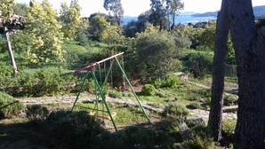 A playground with swings and a climbing structure in a natural setting with trees and a view of the sea in the background. - Spacious ground floor of villa, magnificent sea view, terrace , enclosed parking (Le Lavandou)