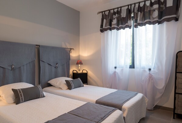 A hotel room with two beds, a nightstand with a lamp, and a window with sheer curtains.