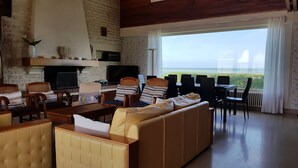 A spacious living area with a sofa, chairs, a coffee table, and a television. There is a fireplace, a large window with a view of the sea, and a dining area with a table and chairs.