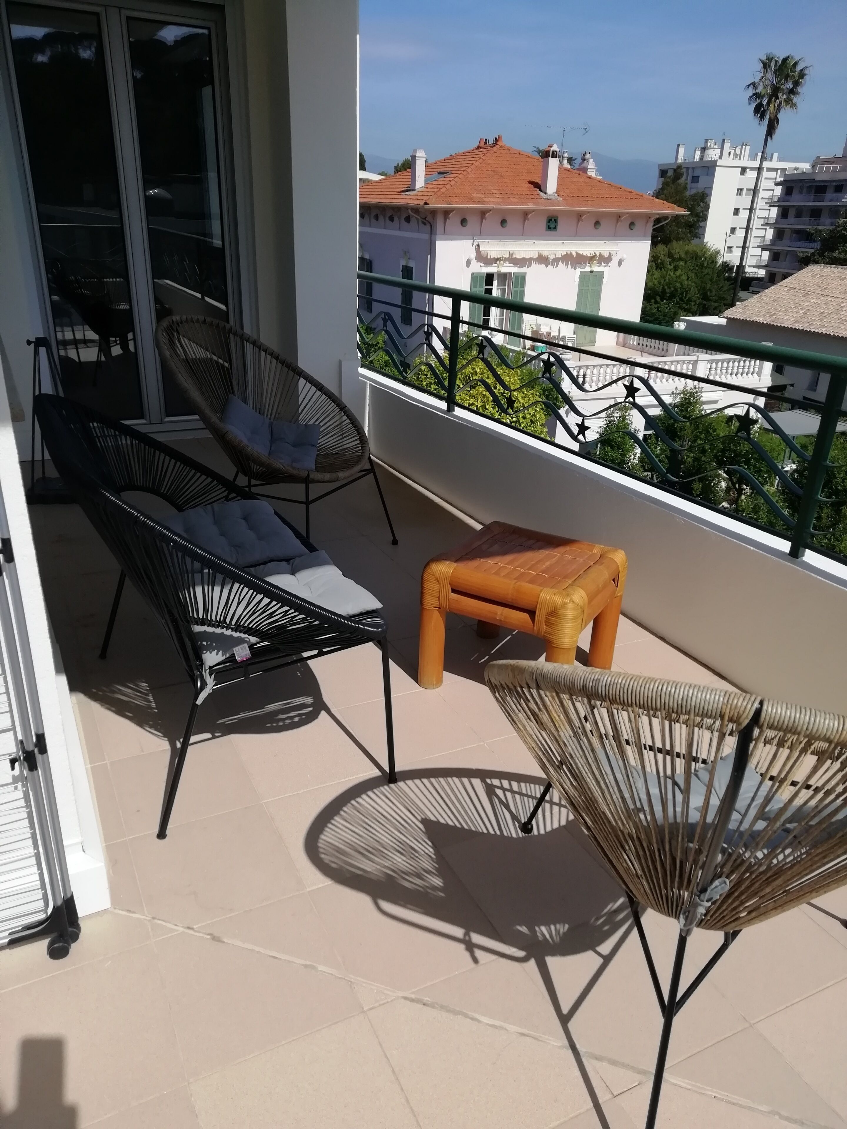 Ein Balkon mit Korbmöbeln, einem kleinen Hocker und Blick auf Gebäude und Bäume.