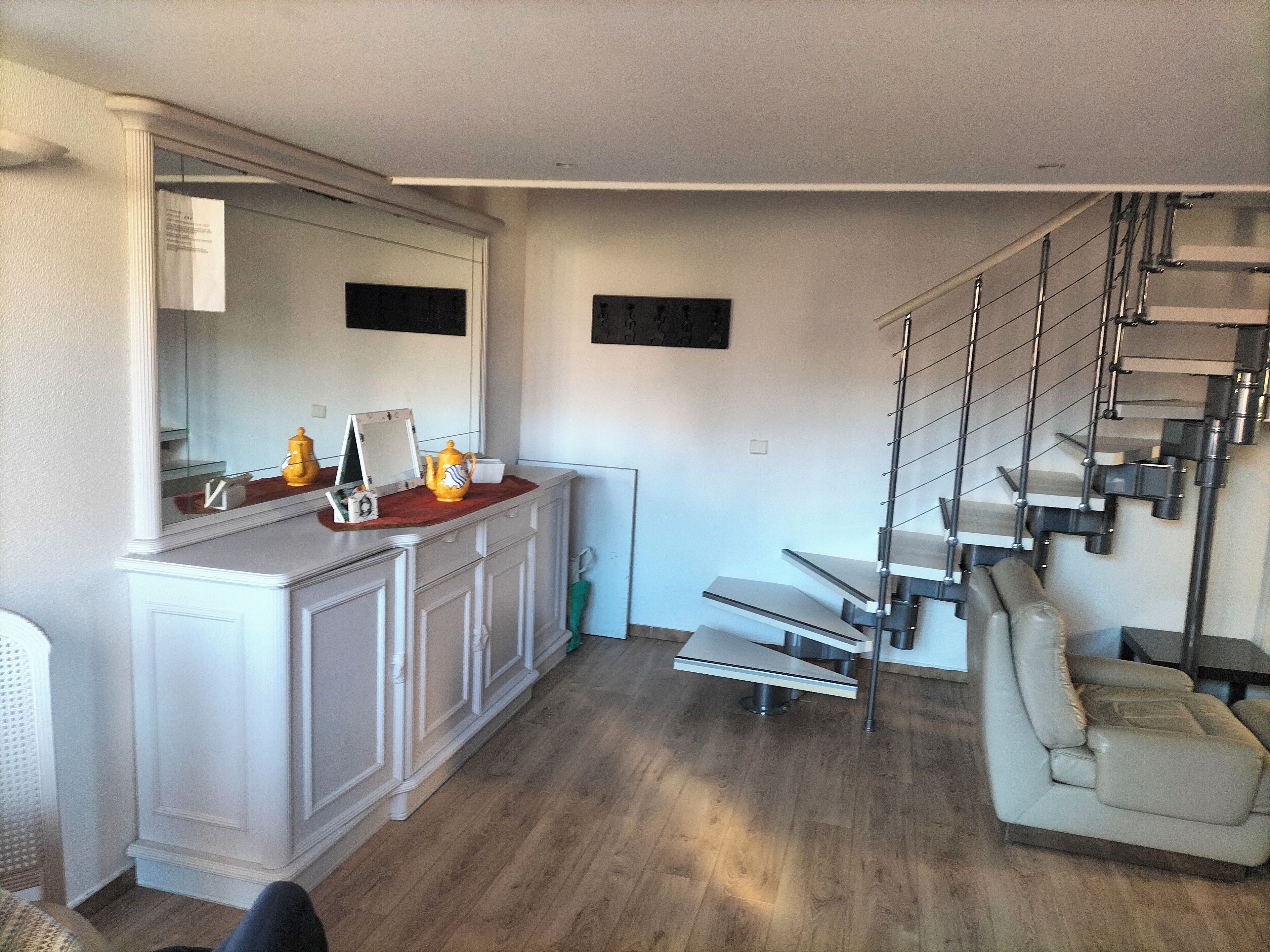 Une cuisine moderne avec des meubles blancs, un plan de travail en bois et un petit îlot central. Un escalier avec une rampe en métal mène à un niveau inférieur.