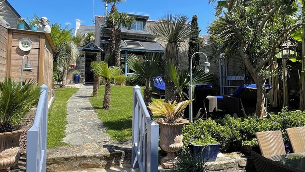 Een pad van stenen leidt naar een huis met een overdekte patio, omringd door potplanten en tuinmeubilair.