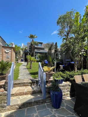 Un chemin de pierres mène à une maison dotée d’un patio couvert, entouré de plantes en pot et de mobilier d’extérieur.