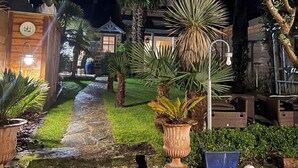 Een tuin 's nachts, met een pad van stenen, verschillende planten in potten en een houten constructie met een lamp.
