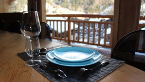 Een eettafel gedekt met een glas, een blauw bord, een vork en een lepel, met zicht op een houten terras en bergen op de achtergrond.