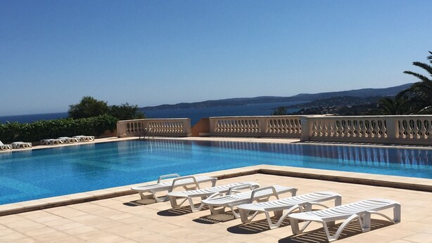 Une piscine bordée de chaises longues blanches, offrant une vue sur la mer et les collines.