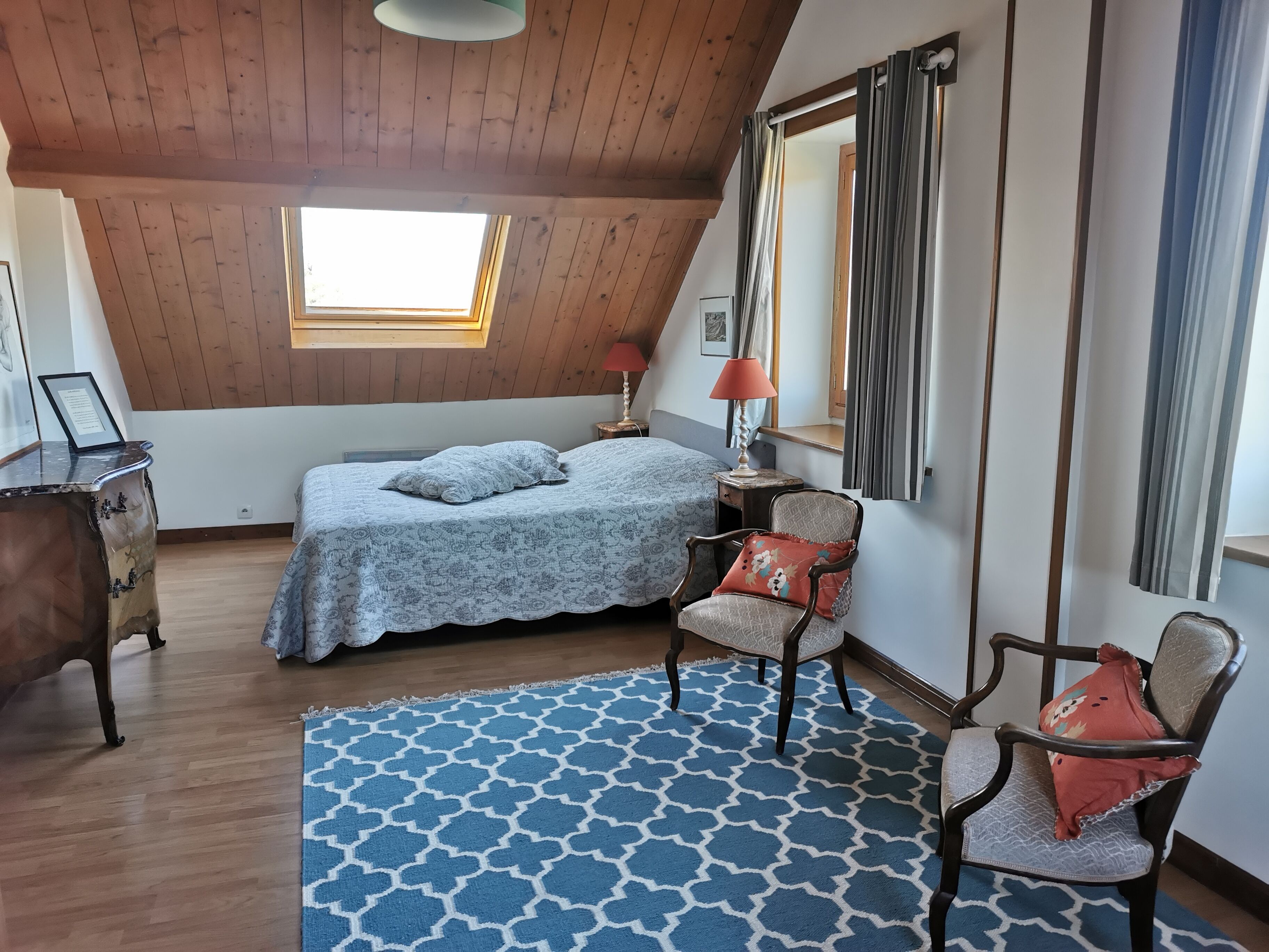 A bedroom with a sloped ceiling, a bed with patterned bedding, a chair with a cushion, a small table with a lamp, and a window with curtains.