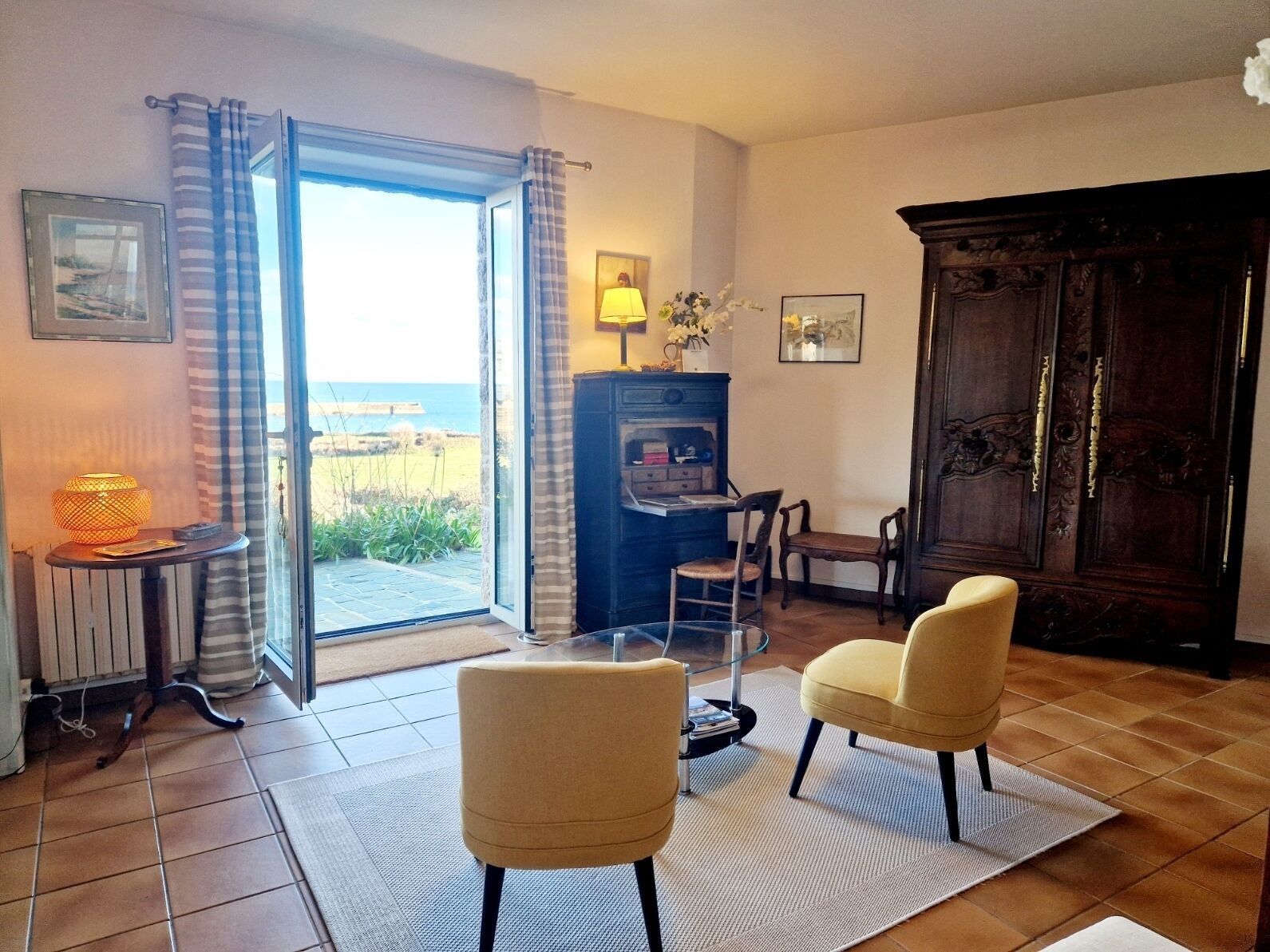 Un salon doté d’une porte vitrée donnant sur un balcon, d’un meuble en bois, de deux fauteuils, d’une petite table avec une lampe et d’une cheminée.