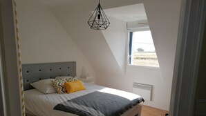 A bedroom with a sloped ceiling, a bed with a grey headboard, a window with a view of the outdoors, and a hanging light fixture. - 3-star seaside villa, sleeps 7, 150 m from the beach and the GR34 trail. Large, enclosed garden (Plounéour-Brignogan-Plages)