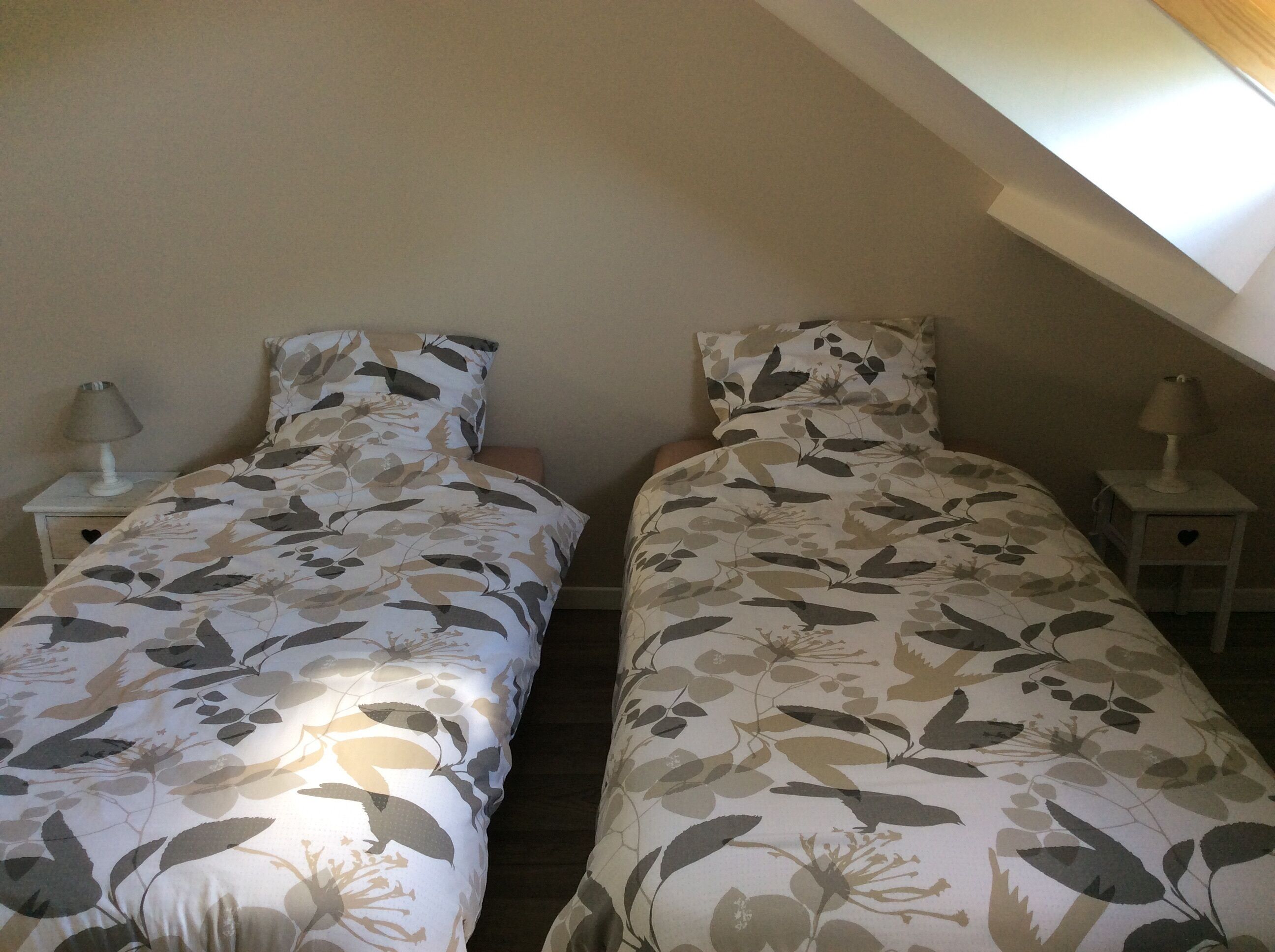 A small, dimly lit room with two single beds, each with a floral-patterned bedspread and matching pillows. There are two bedside tables, each with a lamp. The room has a sloped ceiling, suggesting it is part of an attic or loft space.