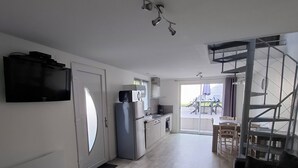 A modern living room with a sofa, a round coffee table with remotes, a kitchen area with a refrigerator, and a staircase leading to an outdoor area.