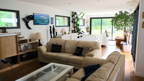 A living room with a sofa, a coffee table, and a bookshelf. There is a large window with a view of greenery outside. - The forest, 400 m from an arm of the sea.  (Carnac)