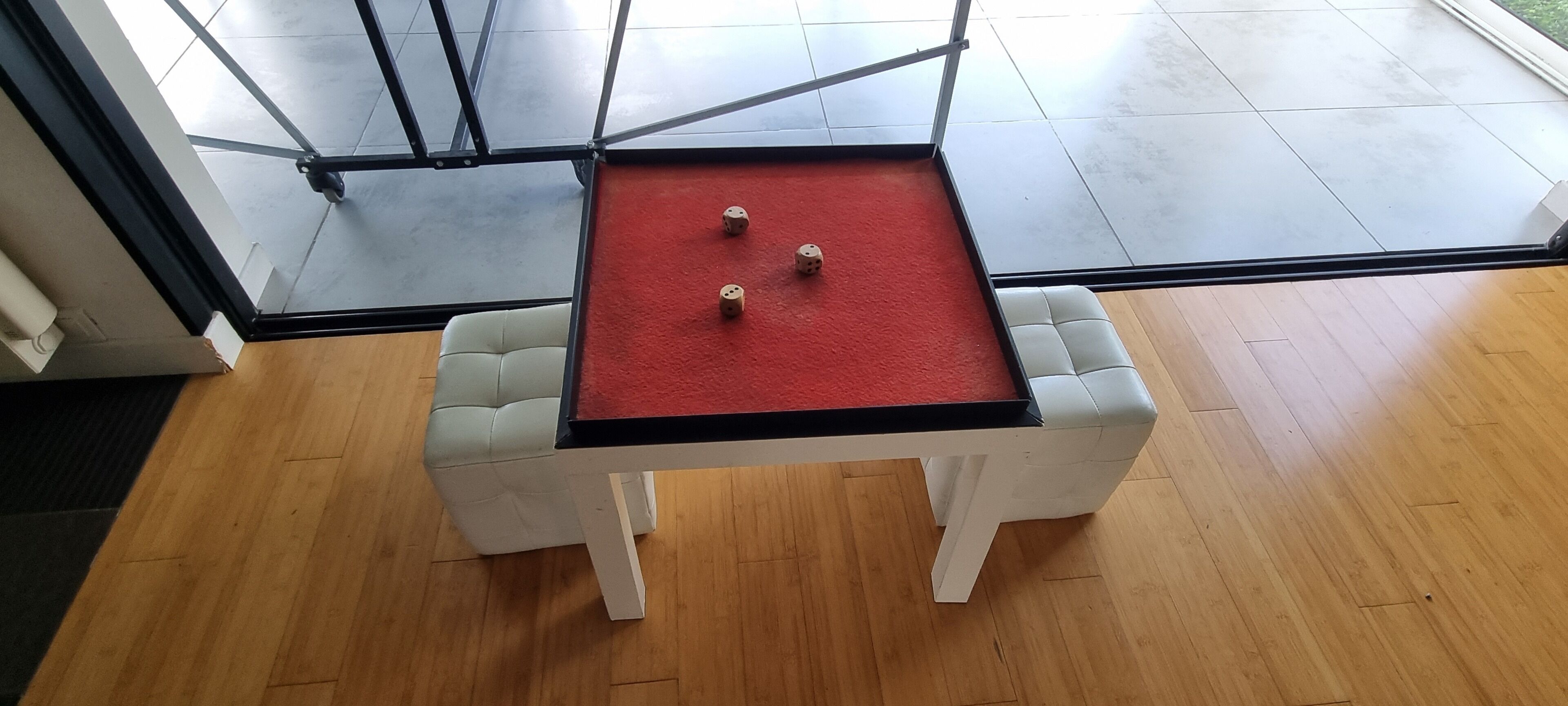 A small table with a red surface and two dice, surrounded by white chairs, in a room with wooden flooring and large windows.