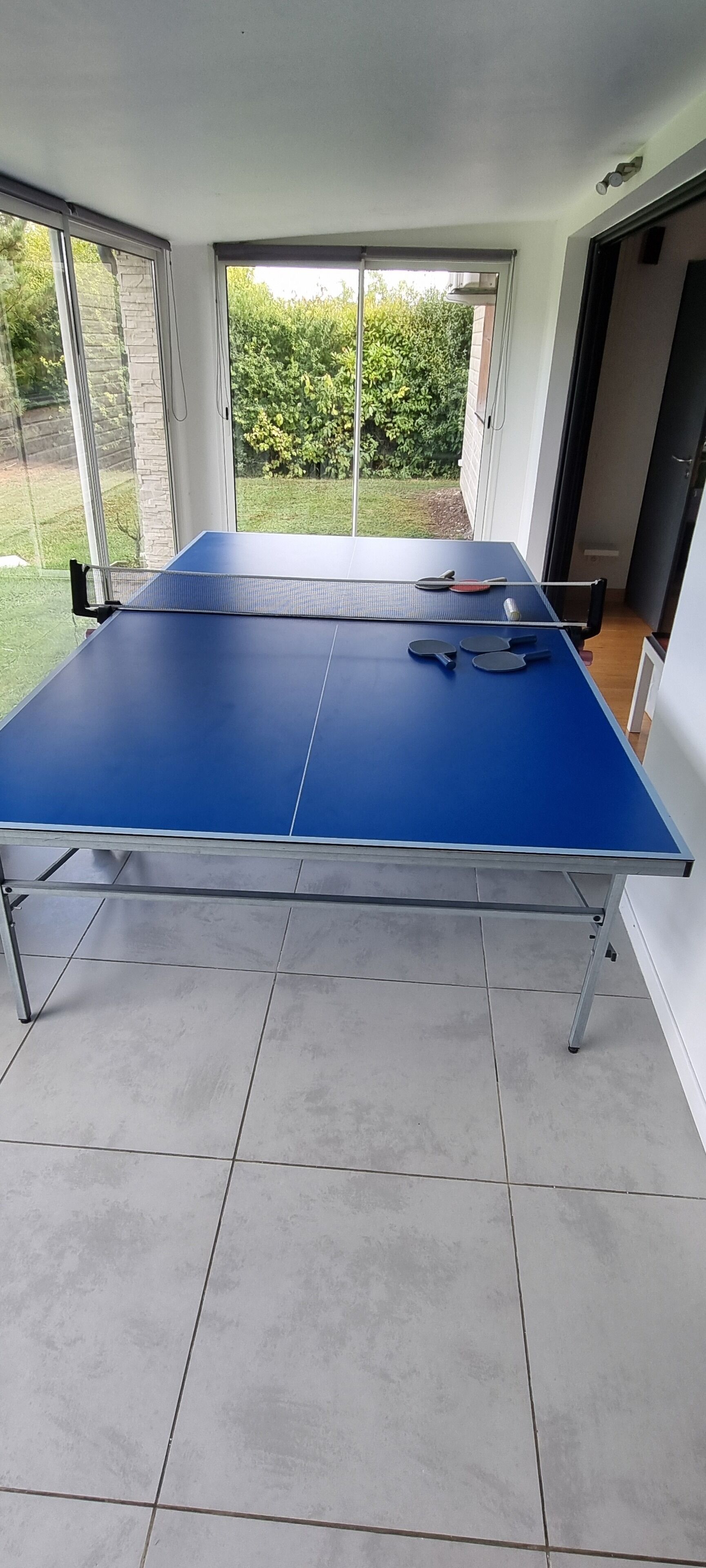 Ein Tischtennis-Tisch mit Schlägern und Bällen in einem Raum mit großen Fenstern und Fliesenboden.