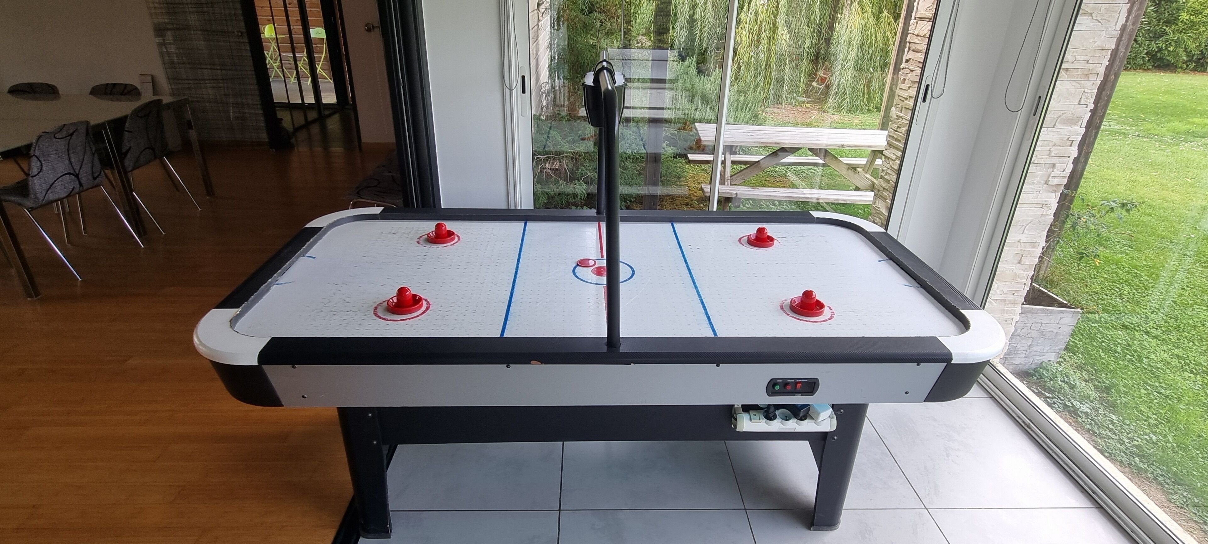 A pool table with four red puck holders, a control panel, and a view of a patio area with a wooden table and chairs.