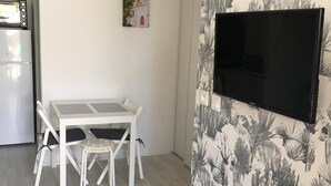 A modern kitchen with white cabinetry, a built-in oven, and a microwave. There is a dining area with a white table and chairs, and a wall-mounted TV. - Cottage in the heart of the pines (Lacanau Océan)