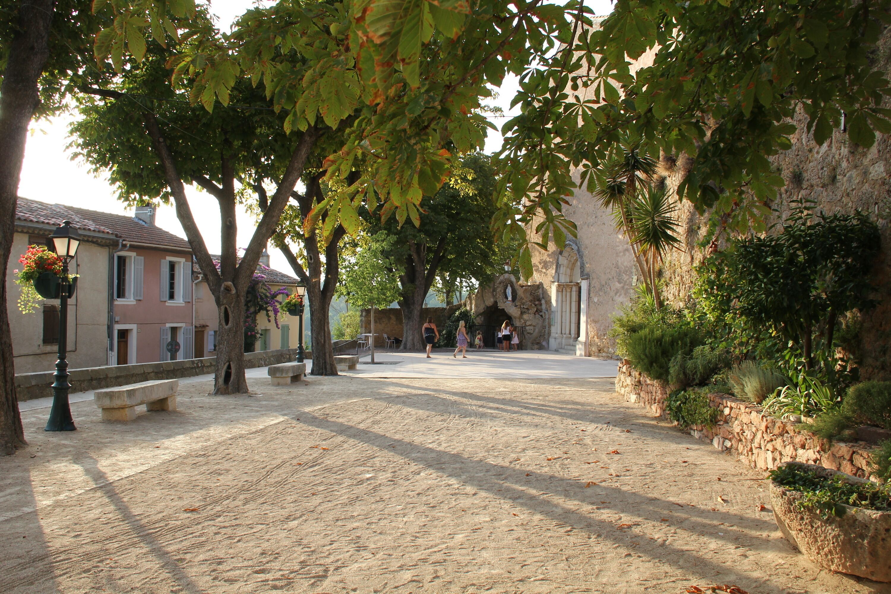 Una plaza adoquinada con bancos y árboles, rodeada de edificios de paredes de piedra y balcones.