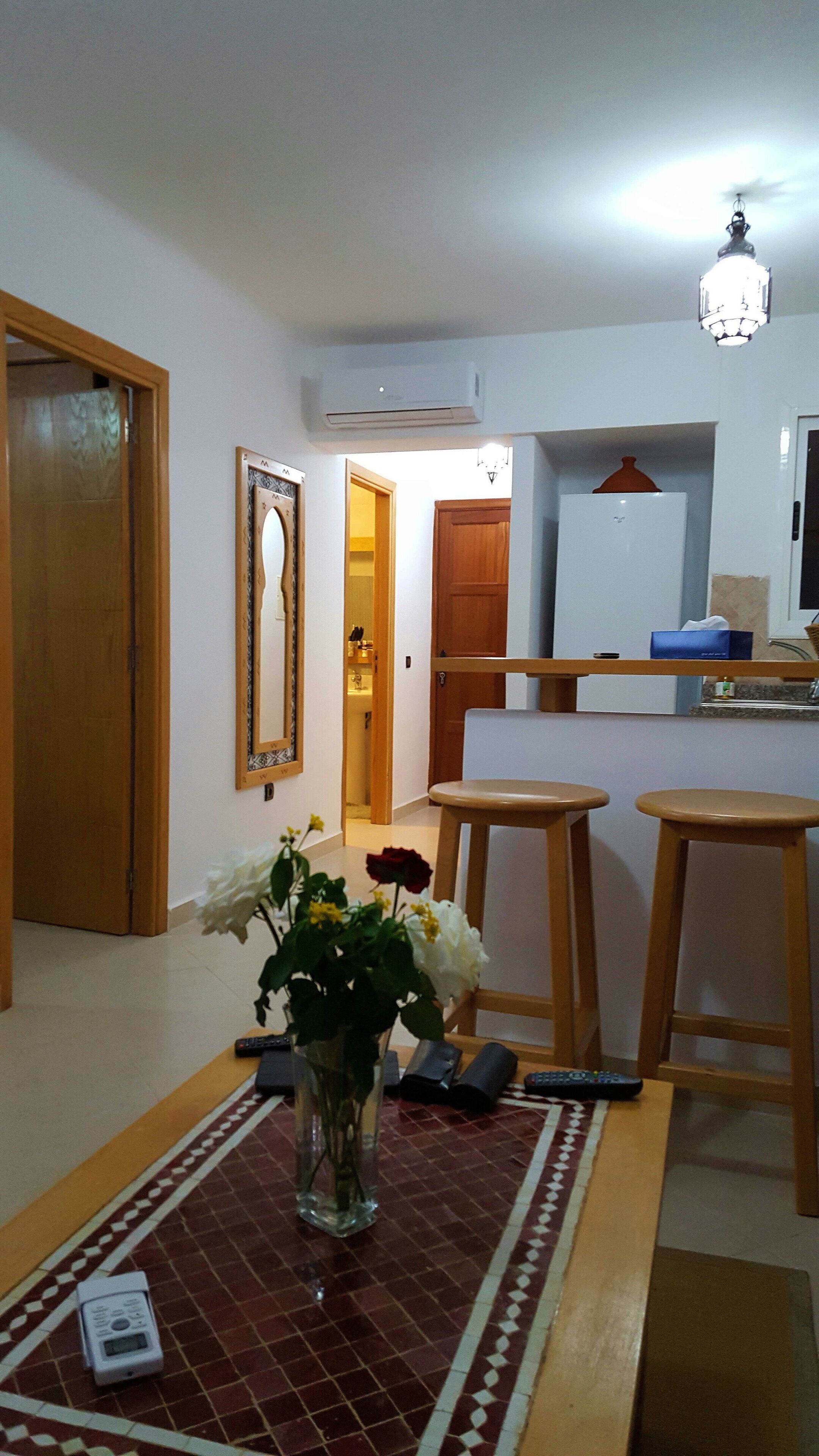 A living room with a wooden floor, a dining table with two stools, a vase with flowers, and a remote control on the table.