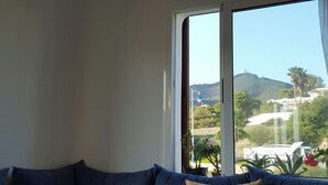 A room with a blue sofa, a vase with white flowers, and a view of a landscape with hills and a white building.