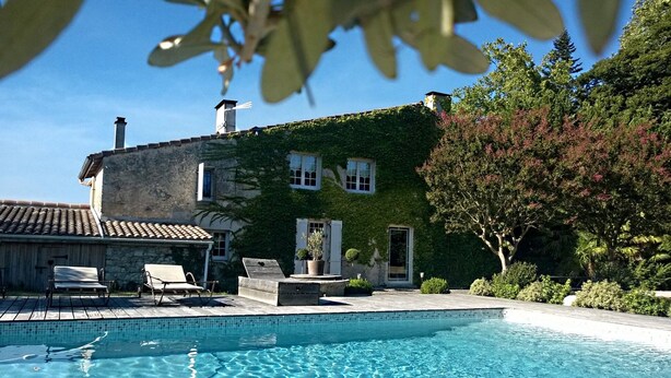 A clear swimming pool with a wooden deck, a stone building with ivy, and a tree with pink flowers.