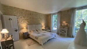 A bedroom with a stone wall, a bed with white bedding, a chair, a table, and a dress on the floor.